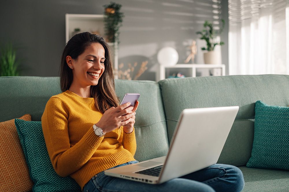 woman using phone and laptop sitting on her couch